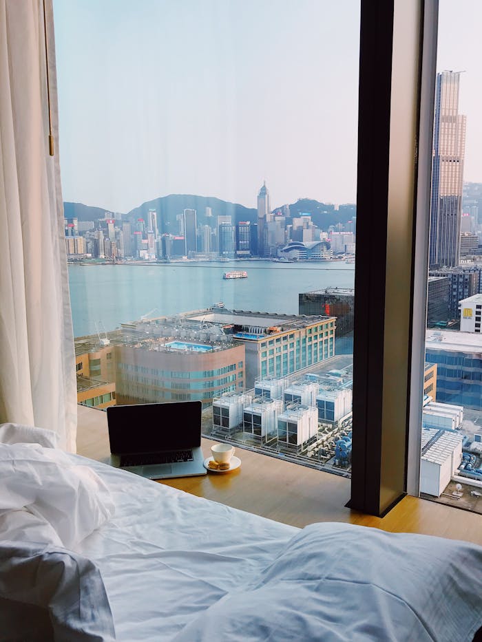 about-01 Cozy hotel room with cityscape view from bed, featuring a laptop and coffee by the window.